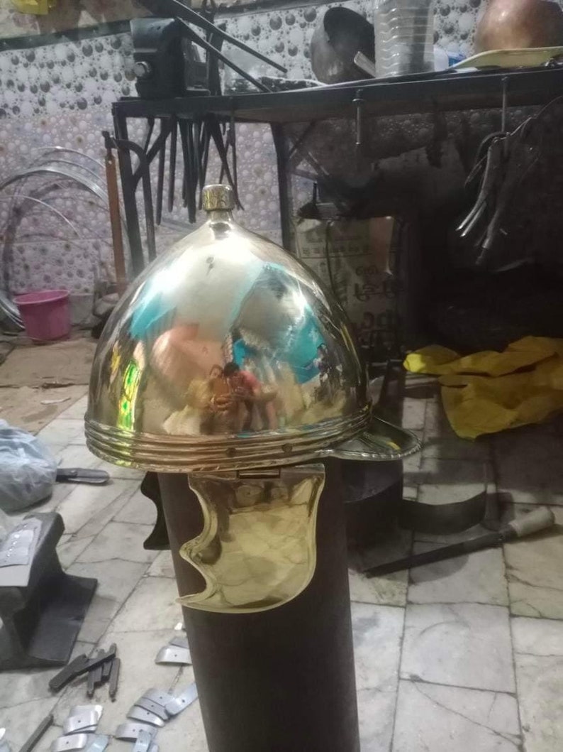 May include: A gold Roman helmet with a hinged cheek guard. The helmet is made of metal and has a shiny finish. It is displayed on a wooden stand.