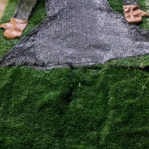 May include: A long-sleeved chainmail hauberk constructed from interlocking silver-toned metal rings, featuring a hood and tan leather cuffs at the end of each sleeve. The garment is laid flat, displaying a flared hem and a protective hood structure.