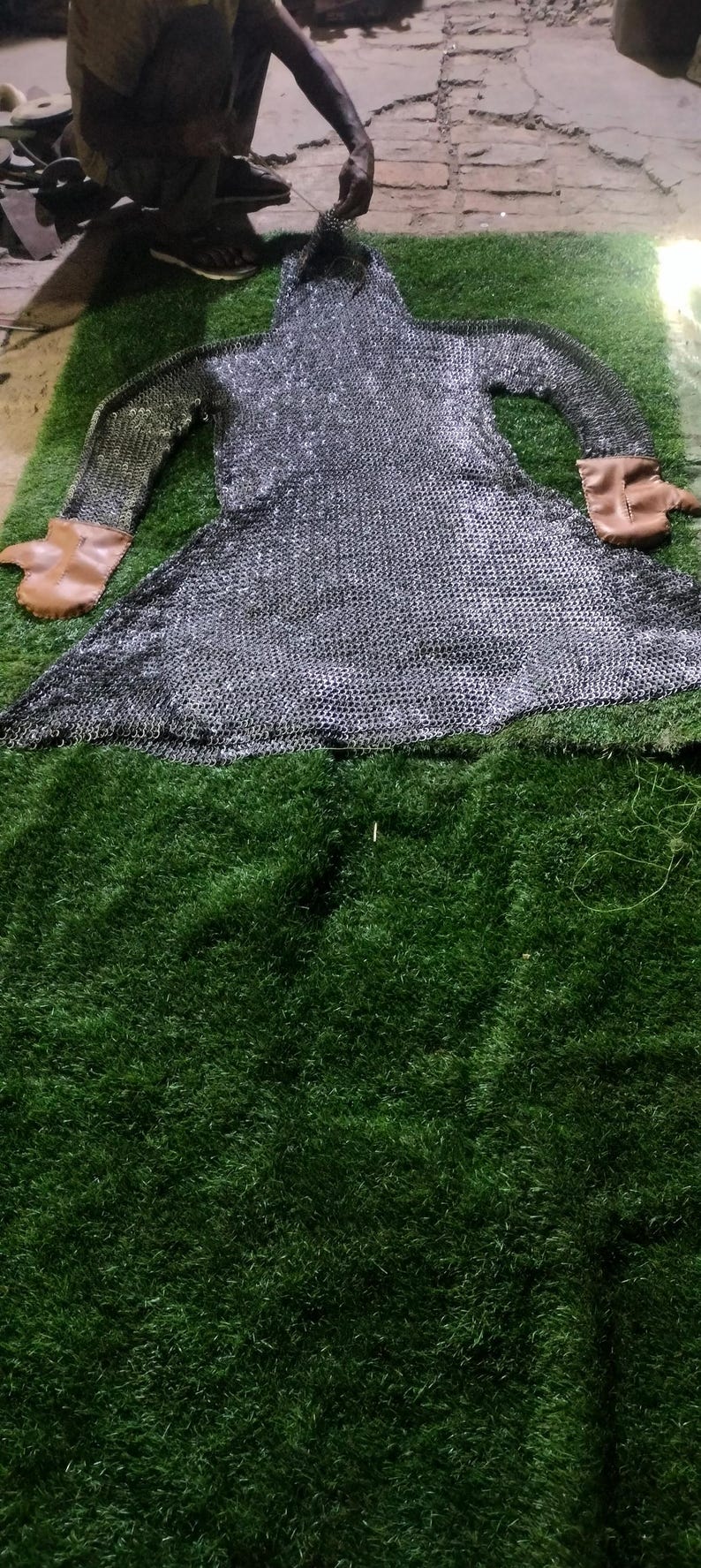 May include: A long-sleeved chainmail hauberk constructed from interlocking silver-toned metal rings, featuring a hood and tan leather cuffs at the end of each sleeve. The garment is laid flat, displaying a flared hem and a protective hood structure.