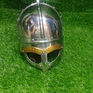 May include: A silver helmet with a gold trim and a pointed top. The helmet is on a green grassy surface.
