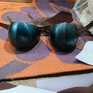 Puede incluir: Dos rodilleras de metal azul turquesa con correas de cuero marrón. Las rodilleras están sobre una alfombra naranja y morada con estampado.
