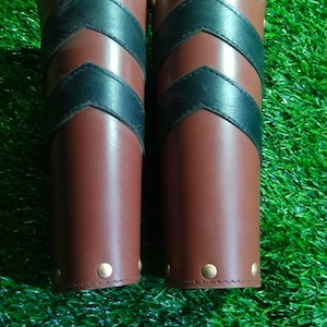 May include: A pair of brown leather arm guards with black leather accents and brass studs. The guards have a chevron pattern.