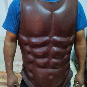 May include: A brown leather chest piece with a sculpted muscle design. The chest piece is worn by a person and has a strap across the shoulders.