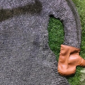 May include: A silver-colored chainmail hauberk featuring a hood and long sleeves. The garment is constructed from interlocking metal rings and includes a tan leather mitten attached to the end of one sleeve, featuring visible stitching along the side.