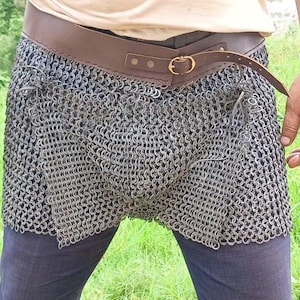 Puede incluir: Una cota de malla plateada que se lleva alrededor de la cintura y los muslos. Se sujeta con un cinturón de cuero marrón con una hebilla dorada. Se lleva sobre unos pantalones azul oscuro.