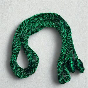 May include: A length of emerald green cord, tightly twisted and looped, lies against a white background. The cord has a subtle sheen, suggesting a silk or satin material. The ends are gathered into small, decorative tassels.