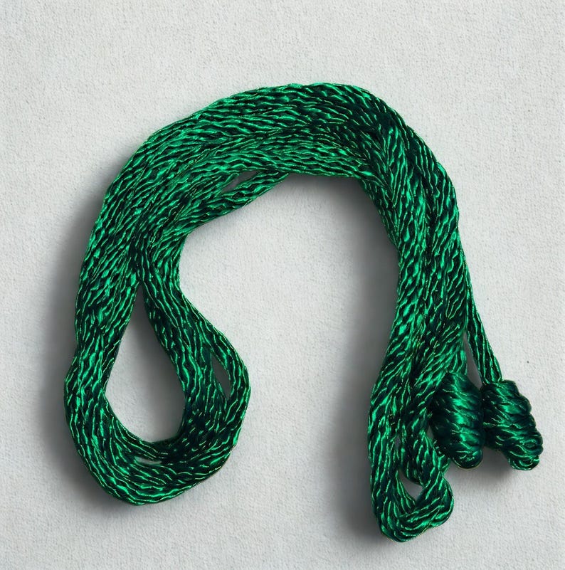May include: A length of emerald green cord, tightly twisted and looped, lies against a white background. The cord has a subtle sheen, suggesting a silk or satin material. The ends are gathered into small, decorative tassels.