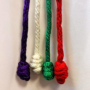 May include: Close-up of four colorful cords: purple, off-white, green, and red. Each cord is tightly braided and knotted at the end. The cords are set against a white background, highlighting their vibrant colors and textures.