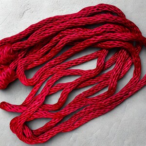 May include: Bright red, twisted cord or rope, possibly yarn, lies in a loose arrangement against a light gray background. The cord appears to be made of a soft, possibly silky material, with a slight sheen. The texture of the cord is visible.