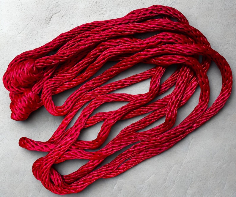 May include: Bright red, twisted cord or rope, possibly yarn, lies in a loose arrangement against a light gray background. The cord appears to be made of a soft, possibly silky material, with a slight sheen. The texture of the cord is visible.