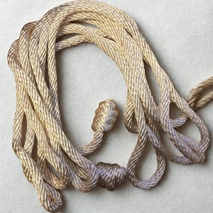 May include: Close-up of a coiled, golden-colored rope with a textured, braided design. The rope has two decorative knots, one at the end of a smaller rope section. The rope is likely used for decorative purposes or crafts.