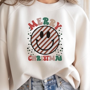 May include: Cream-colored sweatshirt with a "Merry Christmas" design. The design features a smiley face with red and brown stripes, surrounded by green and red text. A red circle with the text "4 SIZES" is in the upper right corner.