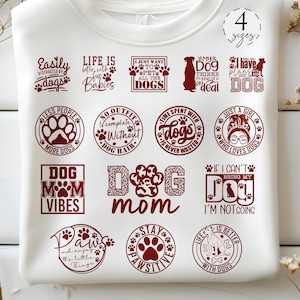 Puede incluir: Una camiseta blanca con 14 diseños diferentes con temática de perros impresos en marrón. Los diseños incluyen frases como "La vida es mejor con bebés de piel", "Mamá de perro", "Solo una chica que ama a los perros" y "Positivo".