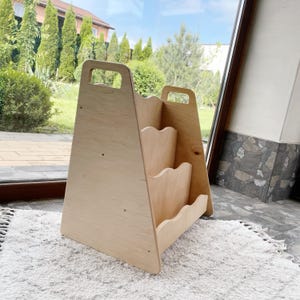 May include: A light-colored wooden bookshelf with a handle at the top and three shelves with a wavy design. The bookshelf is triangular in shape and sits on a white rug.