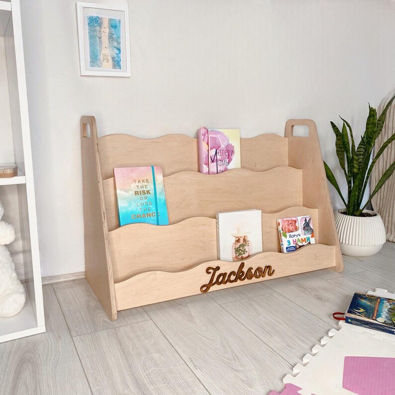 May include: A light-colored wooden bookshelf with three shelves and a wavy design. The name "Jackson" is carved on the front. Several books are displayed on the shelves, with the text "TAKE THE RISK OR LOSE THE CHANCE" visible on one.