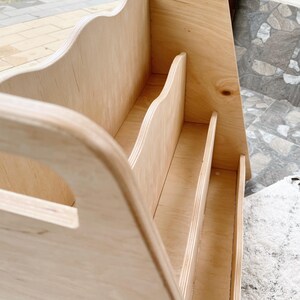 May include: A light-colored wooden organizer with a handle and multiple vertical dividers. The design features curved edges and a natural wood grain finish, suitable for storing books or office supplies.