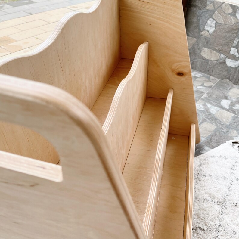 May include: A light-colored wooden organizer with a handle and multiple vertical dividers. The design features curved edges and a natural wood grain finish, suitable for storing books or office supplies.