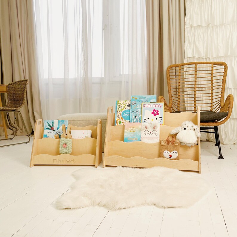 May include: Two wooden bookshelves with a wavy design, filled with children's books and stuffed animals. One shelf has the name "Elizabeth" engraved on it. The shelves are in a bright room with a window and a fluffy white rug.