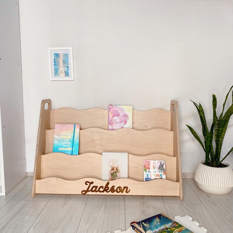 May include: A wooden bookshelf with a wavy design, holding several books. The bookshelf is light-colored and has three shelves. The name "Jackson" is written on the front. A framed picture and a potted plant are in the background.
