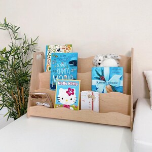May include: A light-colored wooden bookshelf with three shelves, holding several books and a stuffed animal. The books include a Hello Kitty book and a book with a blue cover. A small gift box is also visible.