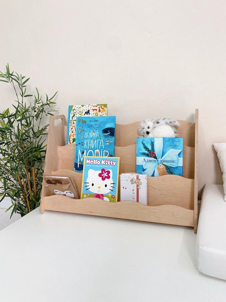 May include: A light-colored wooden bookshelf with three shelves, holding several books and a stuffed animal. The books include a Hello Kitty book and a book with a blue cover. A small gift box is also visible.