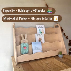 Może przedstawiać: Drewniany regał o minimalistycznym designie, mieszczący kilka książek. Regał ma trzy p&oacute;łki i dekorację w kształcie kr&oacute;lika. Tekst na zdjęciu m&oacute;wi "Pojemność - Mieści do 45 książek" i "Minimalistyczny design - Pasuje do każdego wnętrza."
