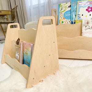 May include: Wooden bookshelf with a handle and a wavy design. The bookshelf is light-colored and holds books and a plush container. The name "Elizabeth" is engraved on the front. A second bookshelf is visible in the background.