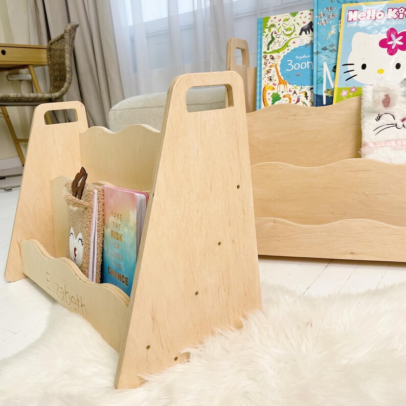 May include: Wooden bookshelf with a handle and a wavy design. The bookshelf is light-colored and holds books and a plush container. The name "Elizabeth" is engraved on the front. A second bookshelf is visible in the background.