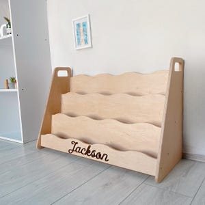 May include: A light-colored wooden bookshelf with a wavy design. The bookshelf has three shelves and a handle on each side. The name "Jackson" is written on the bottom shelf. The bookshelf is made of light-colored wood.