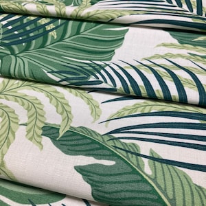 May include: Close-up of a white fabric with a green tropical leaf pattern. The fabric is folded in layers, showing the pattern repeating.