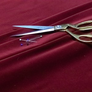 May include: Burgundy velvet fabric with gold-handled scissors and several pins with colored heads. The scissors are open, and the pins are arranged near the blades. The fabric is draped, creating folds and shadows.