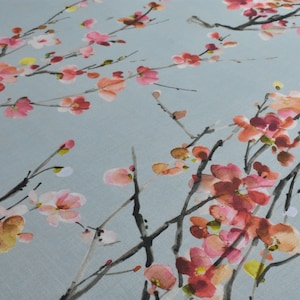 Op de afbeelding: Een lichtblauwe stof met een aquarelprint van roze en rode bloemen op takken. De takken zijn zwart en de bloemen zijn lichtroze, donkerroze en rood.