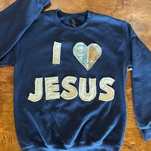 May include: Navy blue sweatshirt with the words "I HEART JESUS" appliqued in off-white fabric. The heart is made of plaid fabric. The sweatshirt is a crew neck style.