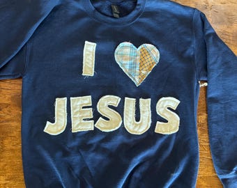 May include: Navy blue sweatshirt with the words "I HEART JESUS" appliqued in off-white fabric. The heart is made of plaid fabric. The sweatshirt is a crew neck style.