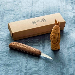 May include: A wooden carving of a Native American person with a headdress, a small wooden knife with a brown wooden handle, and a brown cardboard box with the text "WPC Handcrafted Knives" printed on it.