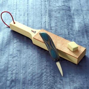 Puede incluir: Un kit de afilado de cuchillos de madera con una correa de cuero, un cuchillo pequeño con un mango azul y gris, y un pequeño bloque de compuesto de afilado blanco. La correa tiene un lazo rojo para colgar.