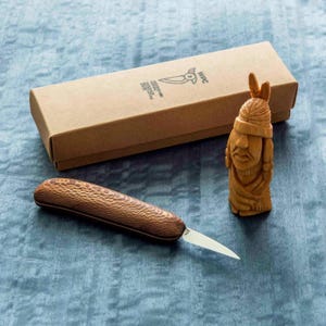May include: A wooden carving knife with a textured handle and a small, sharp blade. Beside it is a carved wooden figure of a Native American and a brown cardboard box with text and a logo.