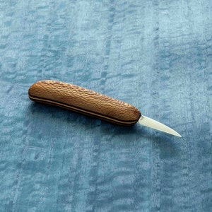 May include: A small, wooden-handled carving knife with a thin, pointed blade. The handle is made of light brown wood with a dark brown pattern.