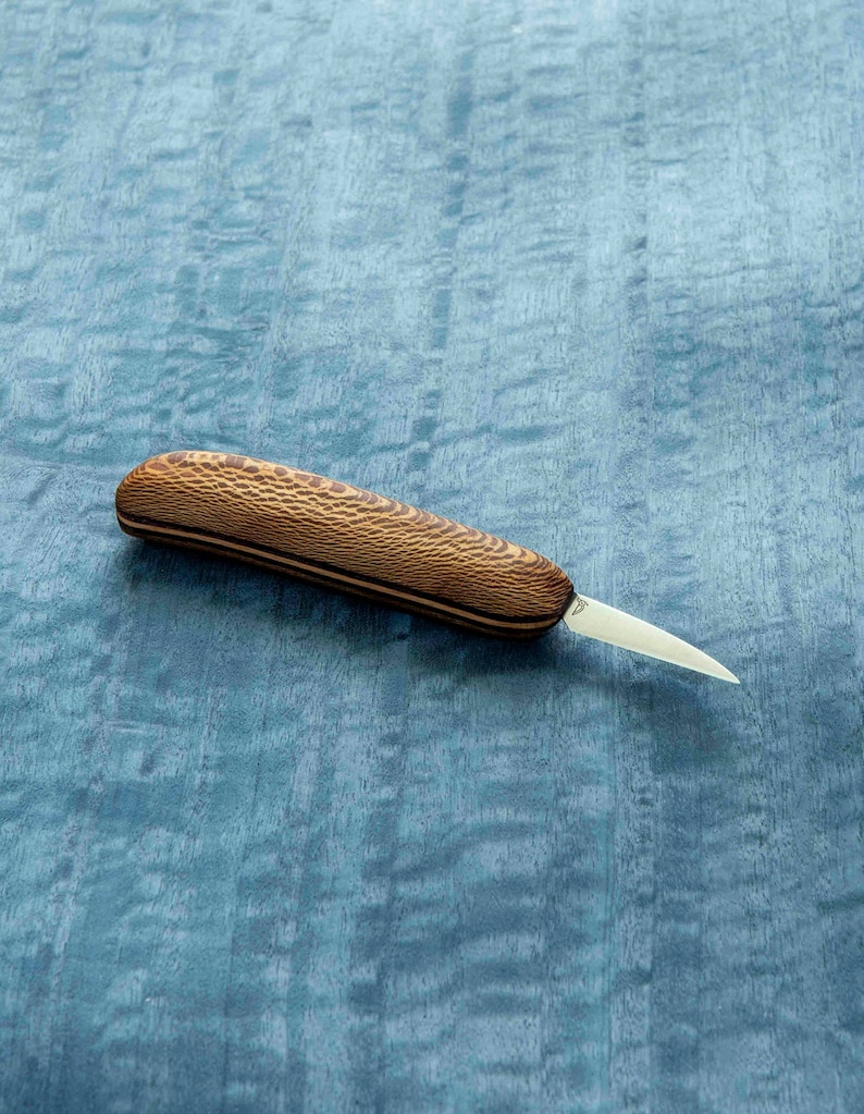 May include: A small, wooden-handled carving knife with a thin, pointed blade. The handle is made of light brown wood with a dark brown pattern.