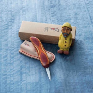 May include: A wooden knife with a red and orange handle, a leather sheath, a cardboard box, and a small carved wooden figurine of a man in a yellow coat and hat. The knife blade is silver.