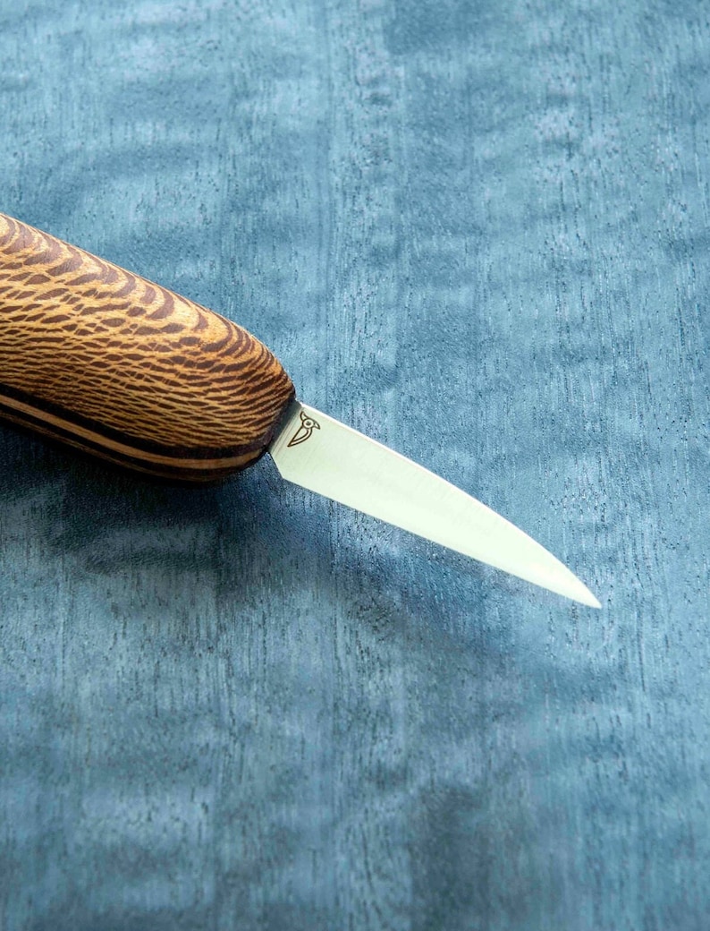 May include: A close-up of a small, wooden-handled knife with a thin, sharp blade. The blade is made of stainless steel and has a slight curve to it. The handle is made of dark wood with a light wood inlay. The knife is lying on a blue surface.