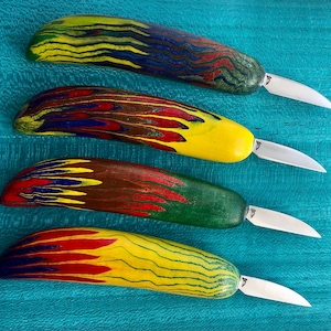 May include: Four small knives with thick, rounded handles made of multi-coloured layered wood. Each handle features distinct swirling patterns in shades of yellow, red, blue, and green. The short, pointed metal blades are attached to the handles.