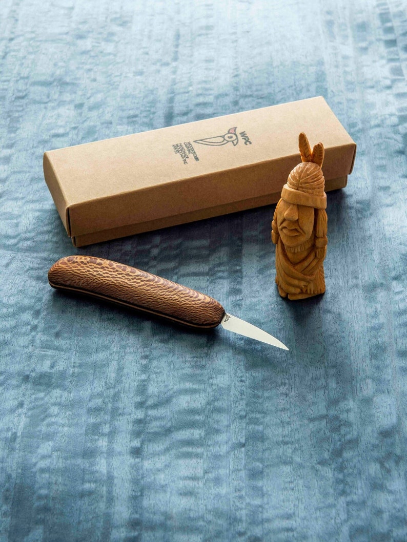 May include: A wooden carving of a Native American person with a headdress, a small wooden knife with a brown wooden handle, and a brown cardboard box with the text "WPC Handcrafted Knives" printed on it.