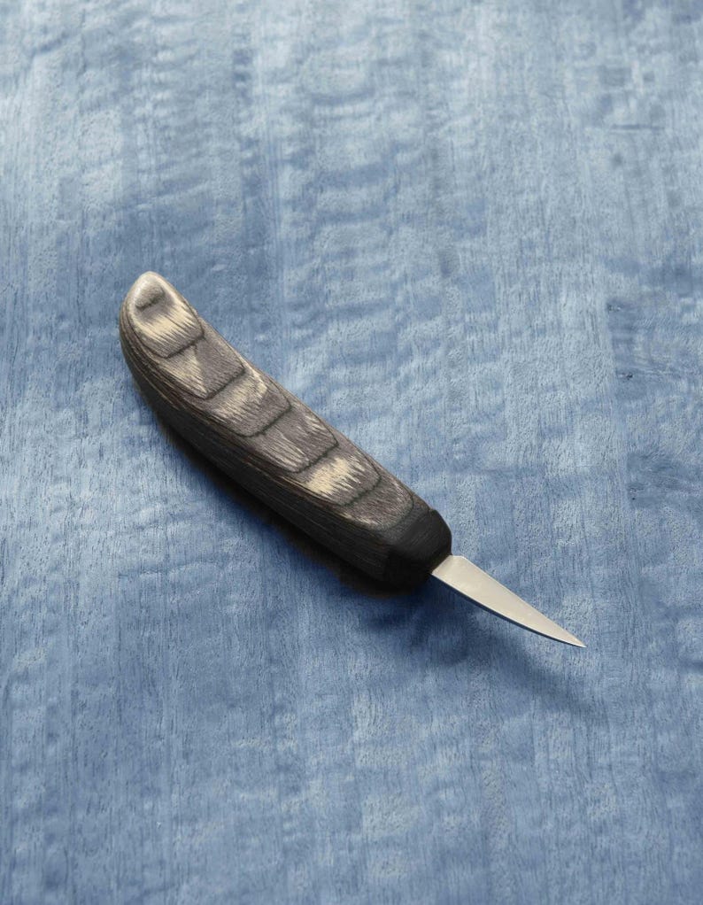 May include: A small, black and grey wood handled carving knife with a thin, sharp blade. The knife is lying on a blue surface.