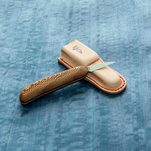 May include: A small, brown handled knife with a silver blade is resting in a brown leather sheath with red stitching. The sheath has the word "WPC" embossed on it.