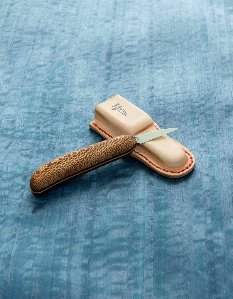 May include: A small, brown handled knife with a silver blade is resting in a brown leather sheath with red stitching. The sheath has the word "WPC" embossed on it.