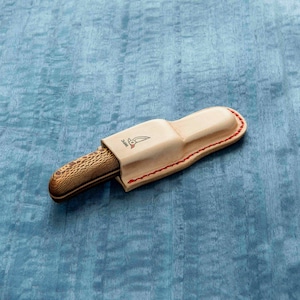 May include: A brown and tan pocket knife with a wooden handle and a leather sheath. The sheath has a red stitching detail and a small logo on the front.