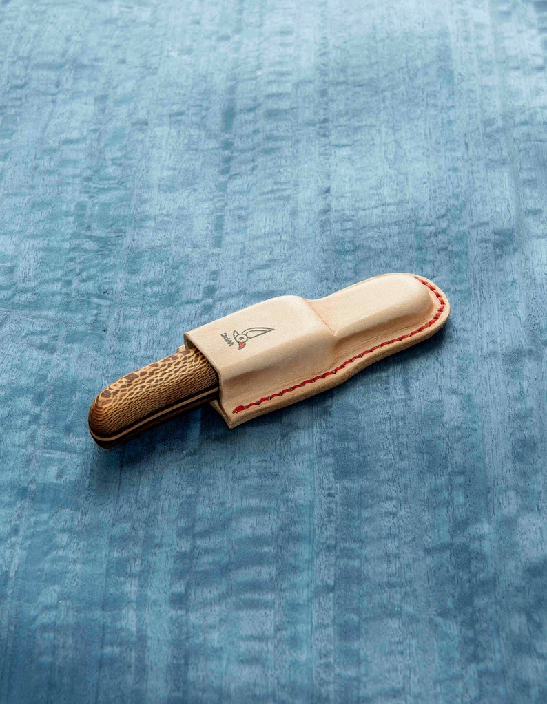 May include: A brown and tan pocket knife with a wooden handle and a leather sheath. The sheath has a red stitching detail and a small logo on the front.