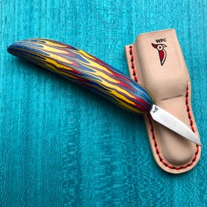 Puede incluir: Un cuchillo artesanal con un mango de madera a rayas de colores azul, amarillo y rojo. El cuchillo tiene una hoja plateada y descansa junto a una funda de cuero color canela con costuras rojas y un logotipo de pájaro. La funda tiene el texto "WPC".