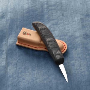 May include: A small, black handled carving knife with a leather sheath. The knife has a curved blade and a wooden handle with a dark wood grain.